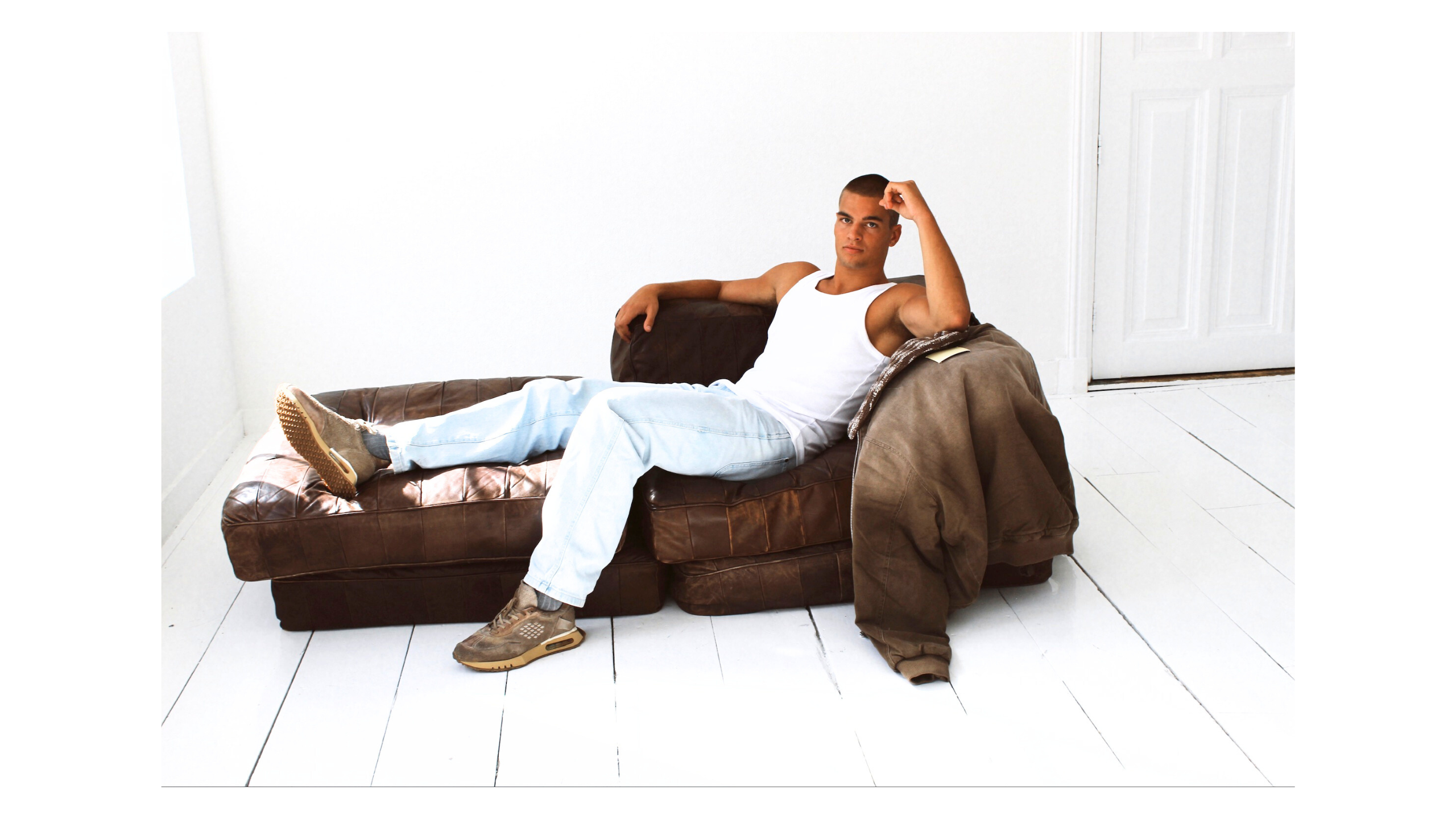 Man sitting on a brown leather sofa in a minimalistic room with white walls and floor. Wearing Bepositive+ sneakers 